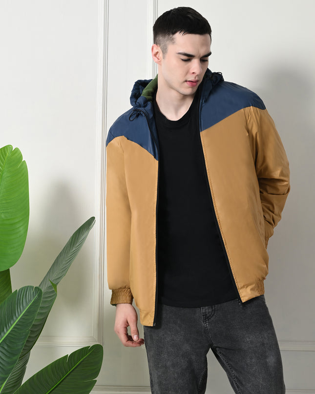 A man wearing a color-blocked windblock jacket with fleece inside mustard yellow and navy blue, with a hood, standing next to a plant.